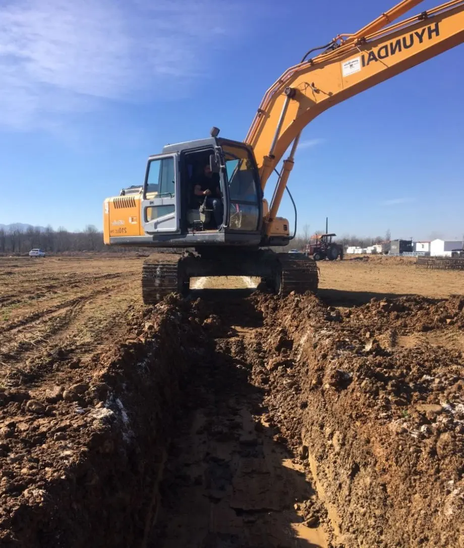 Excavator cutting a foundation trench across a cleared greenhouse site under a clear sky